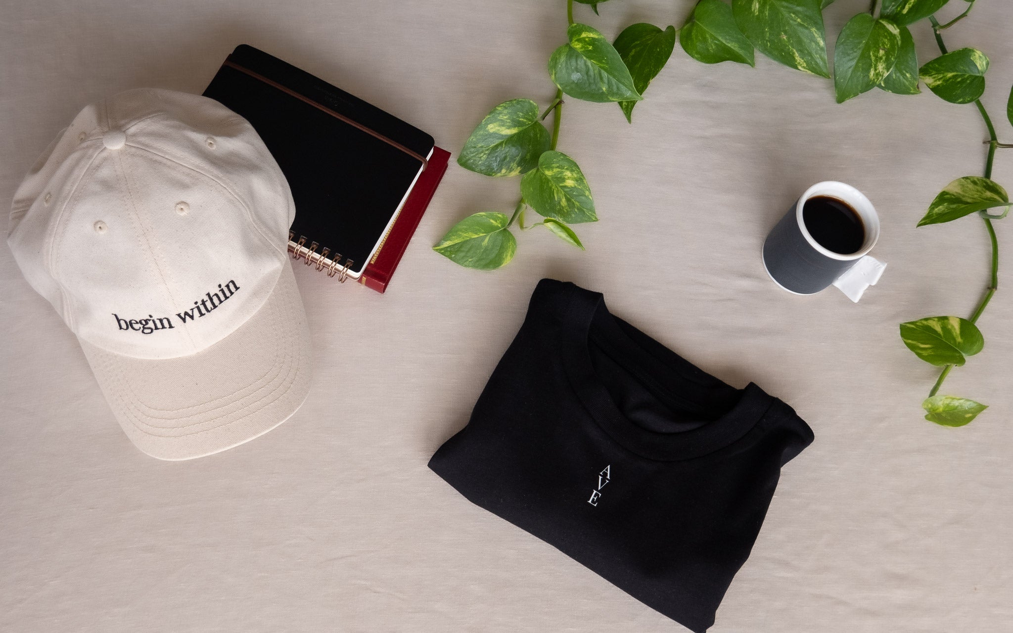 AVE flatlay, begin within dad cap and black TESSERA t-shirt with notebooks, coffee and a plant on natural linen